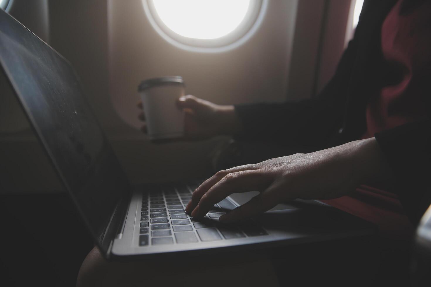 Blonde female tourist checking incoming notification on smartphone sitting on seat of airplane with netbook.Young businesswoman share media from telephone on laptop computer during plane flight photo