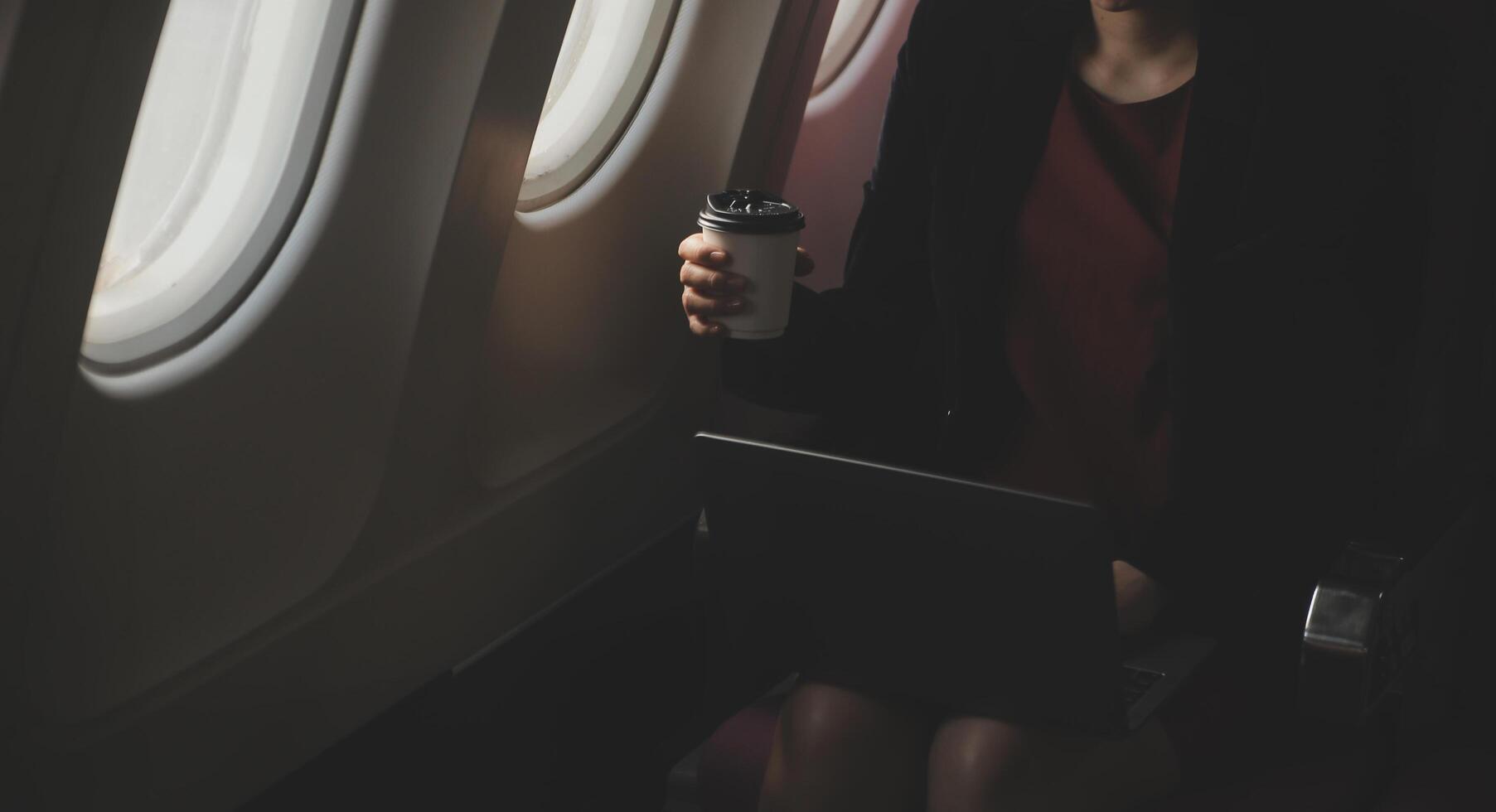 Blonde female tourist checking incoming notification on smartphone sitting on seat of airplane with netbook.Young businesswoman share media from telephone on laptop computer during plane flight photo