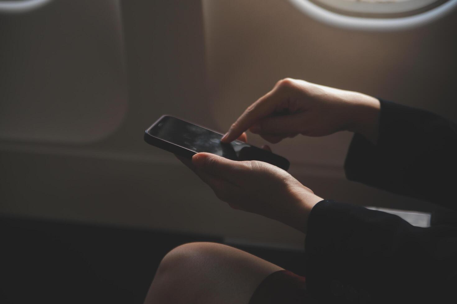 Blonde female tourist checking incoming notification on smartphone sitting on seat of airplane with netbook.Young businesswoman share media from telephone on laptop computer during plane flight photo