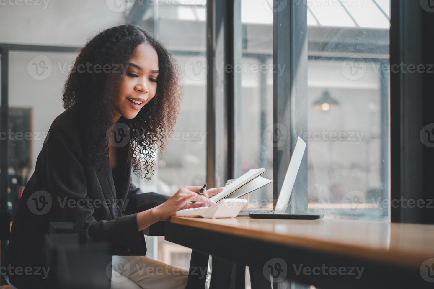 Portrait of attractive hardworking businesswoman with Afro hairstyle busy doing paperwork at office desk, working through finances, using calculator and making notes in her notebook with pen photo