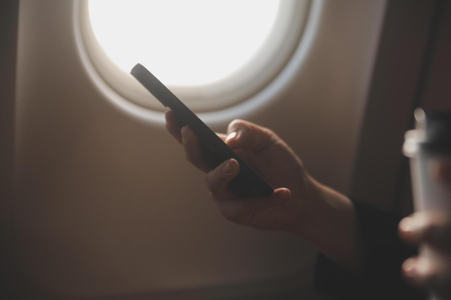 Blonde female tourist checking incoming notification on smartphone sitting on seat of airplane with netbook.Young businesswoman share media from telephone on laptop computer during plane flight photo