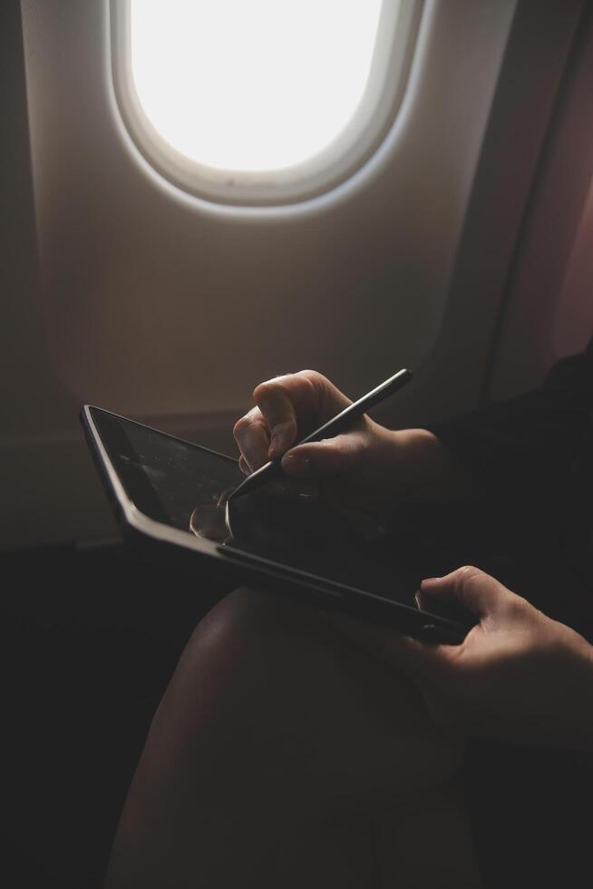 Blonde female tourist checking incoming notification on smartphone sitting on seat of airplane with netbook.Young businesswoman share media from telephone on laptop computer during plane flight photo