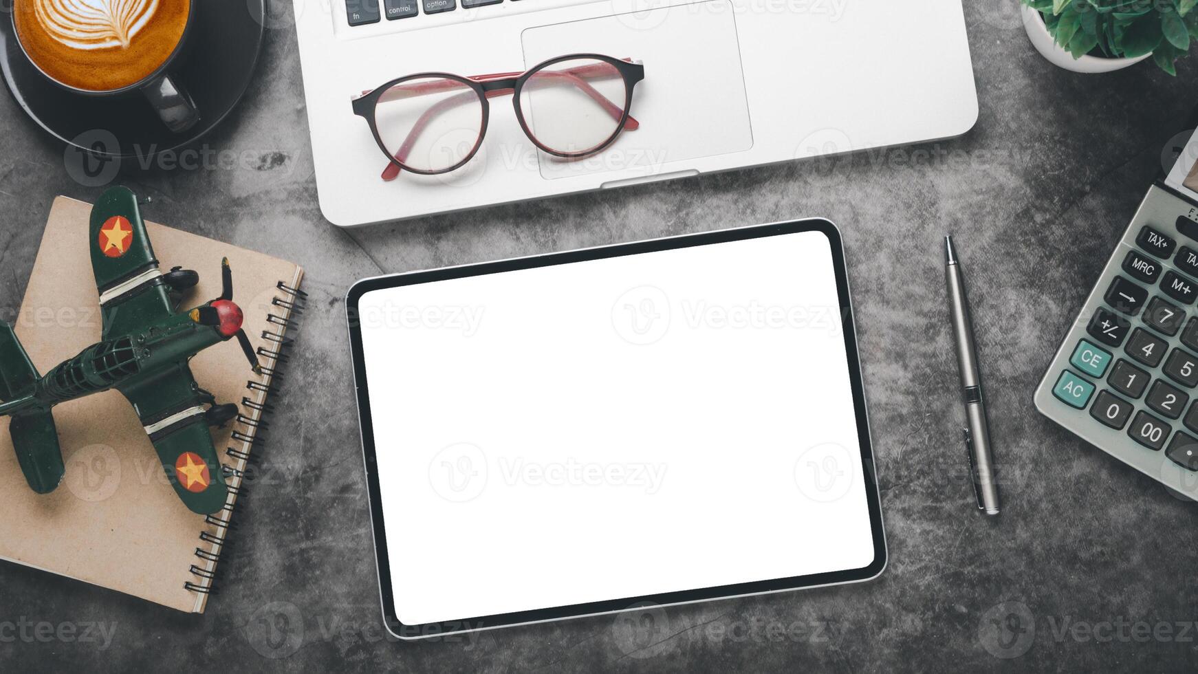 An organized desk layout with a blank tablet screen ready for work, a pair of glasses, a pen, a vintage toy airplane on a notebook, a cup of coffee with latte art, and a calculator. photo