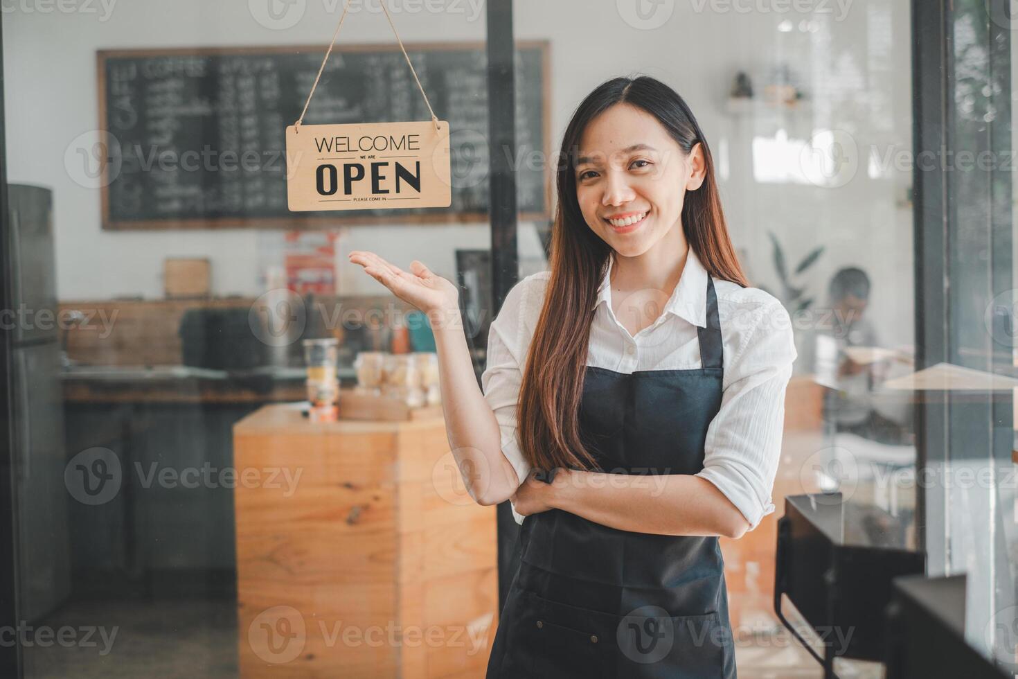 A welcoming cafe owner stands with a bright smile, gesturing to an Open sign, inviting customers into her cozy establishment. photo