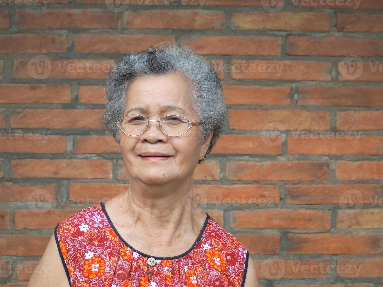 An elderly Asian woman smiling and looking at the camera while standing with a brick wall background. Space for text. Concept of aged people and healthcare photo