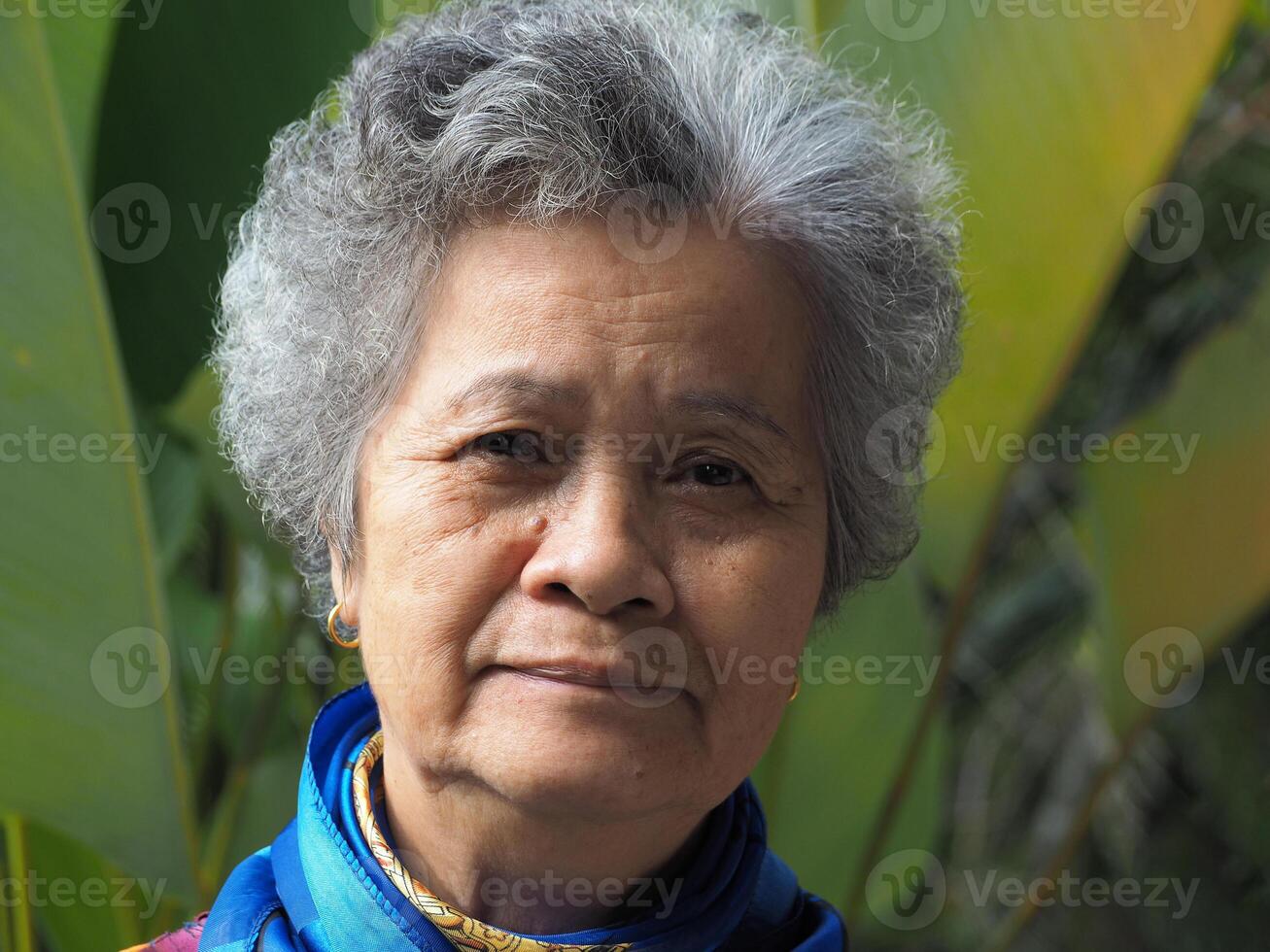 Senior Asian woman with short white hair smiling and looking at the camera while standing in a garden. Space for text. Concept of aged people and healthcare photo