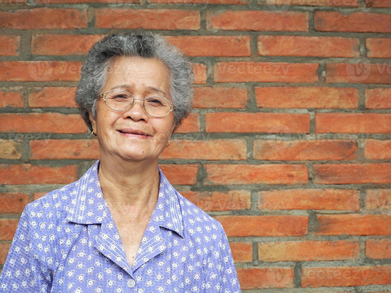 An elderly Asian woman smiling and looking at the camera while standing with a brick wall background. Space for text. Concept of aged people and healthcare photo