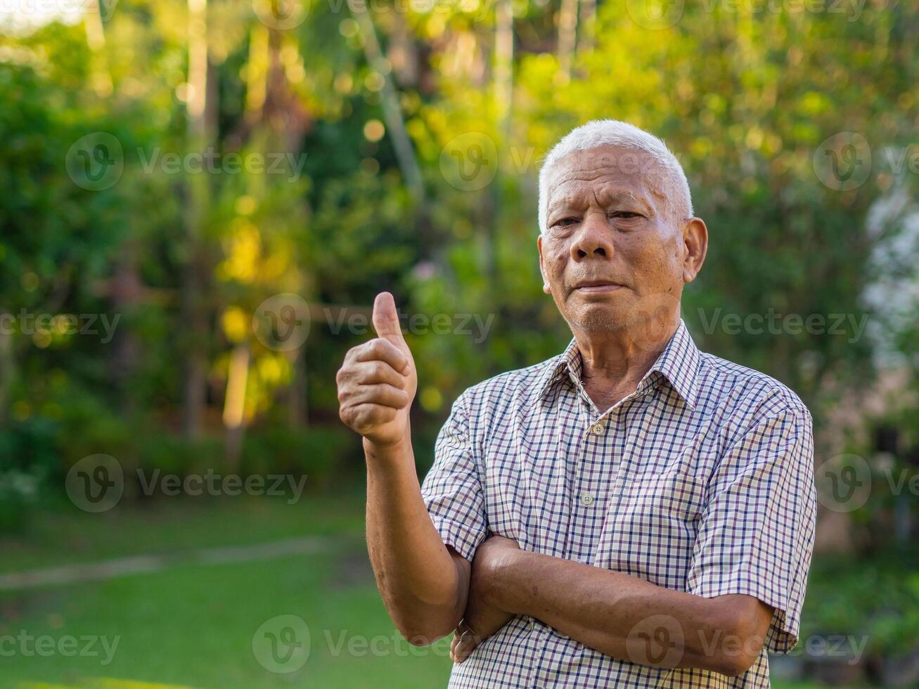 Senior Asian man showing thumb up gesture and looking at the camera while standing in a garden. Space for text. Concept of aged people and healthcare photo