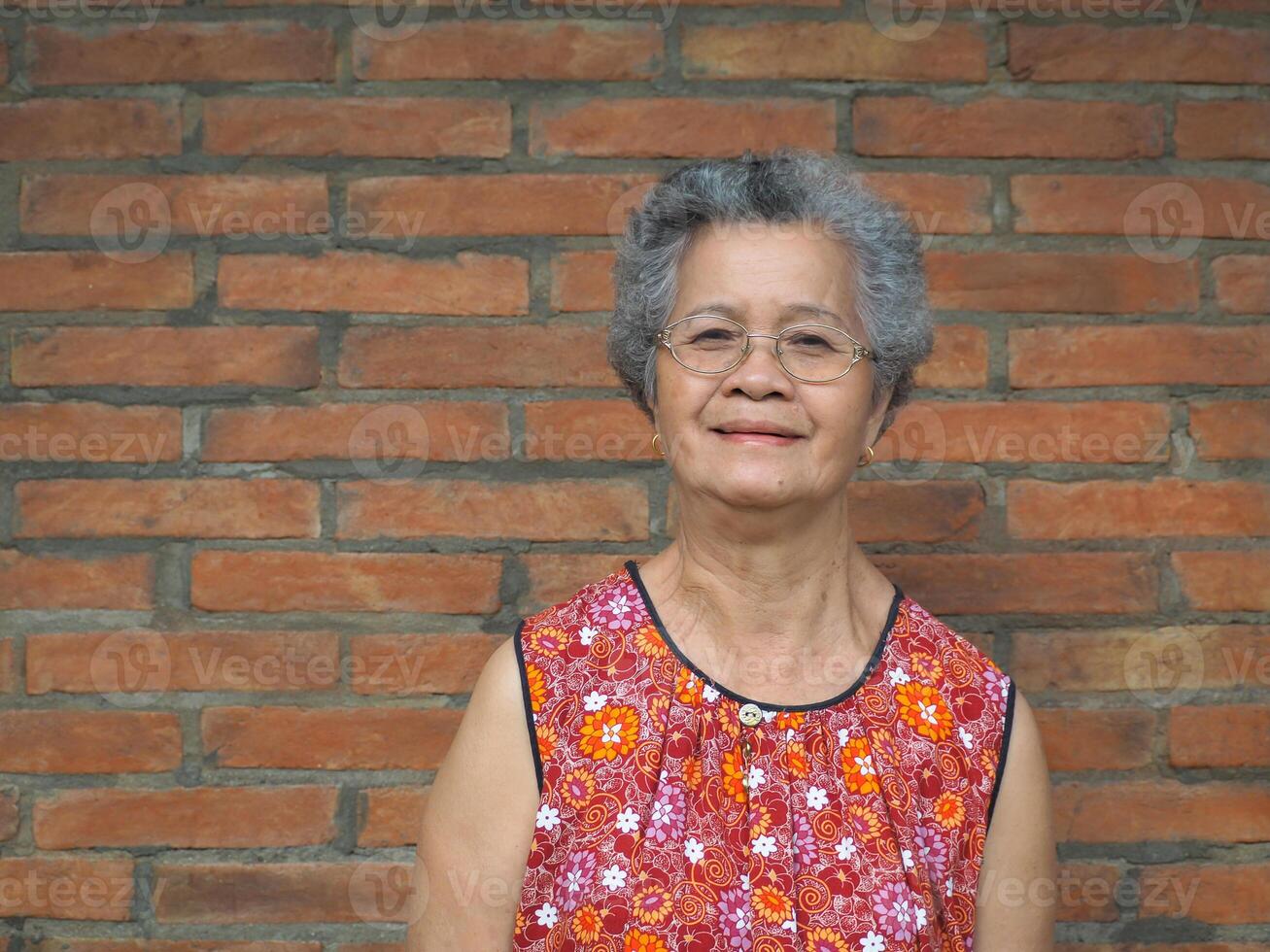 un mayor asiático mujer sonriente y mirando a el cámara mientras en pie con un ladrillo pared antecedentes. espacio para texto. concepto de Envejecido personas y cuidado de la salud foto