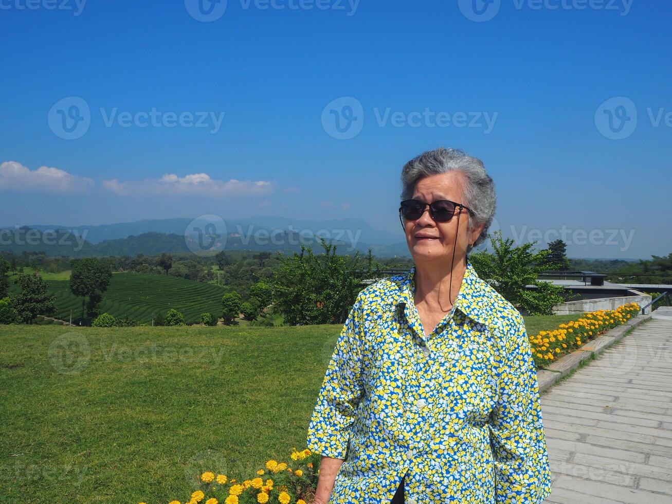 Senior woman wearing sunglasses, smiling and looking at the camera while standing in the tea plantation. Space for text. Concept of aged people and relaxation photo