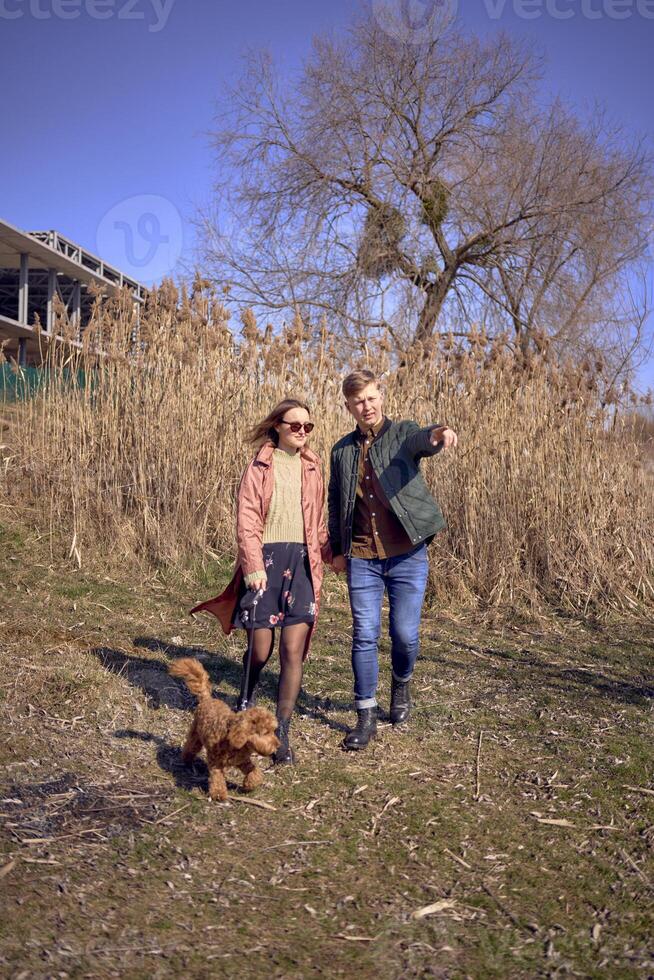 a young stylish couple walking with their cockapoo dog near the river, an unfinished parking lot in the background photo