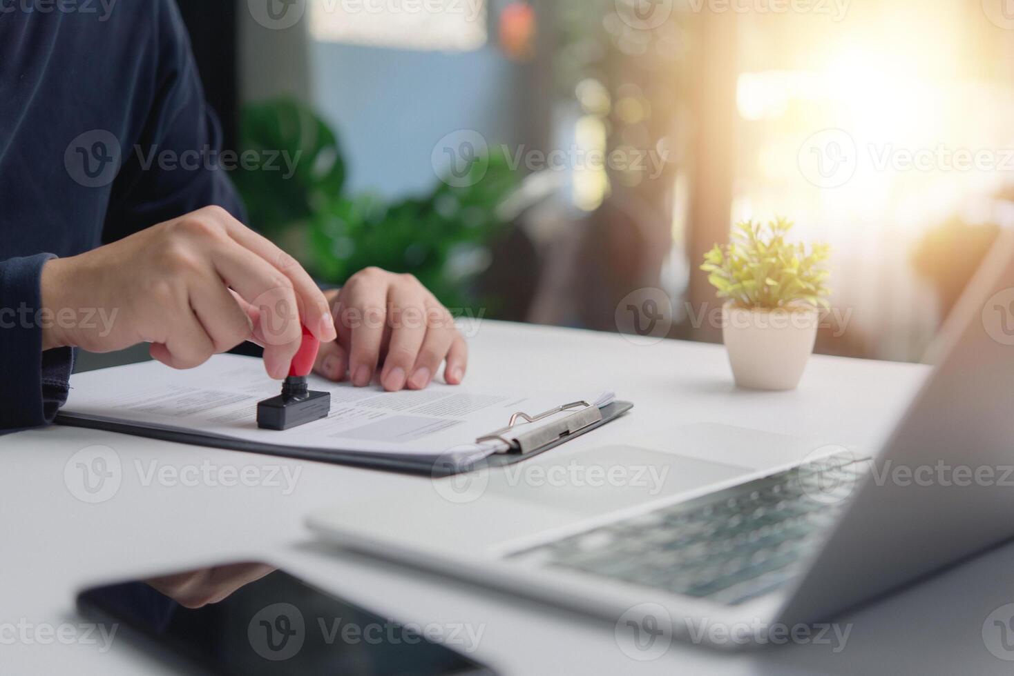 Individual in an office setting applies a stamp to a document contract legal agreement, signifying approval or verification certification permit visa next to an open laptop. photo