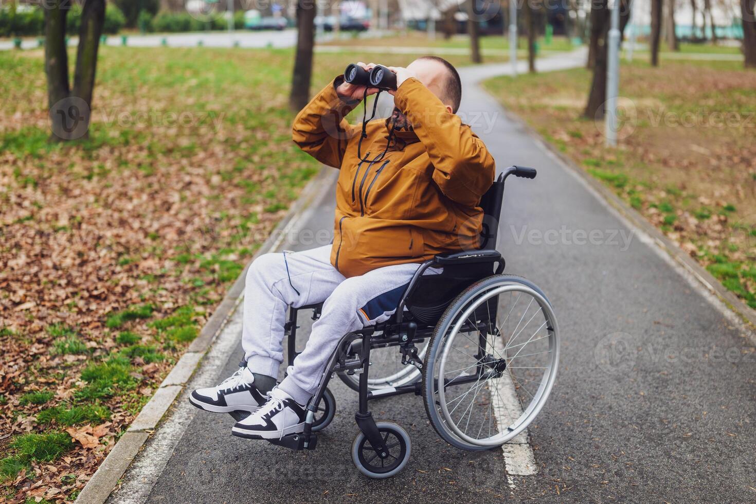 Paraplegic handicapped man in wheelchair is using binoculars outdoor. He is watching birds. photo