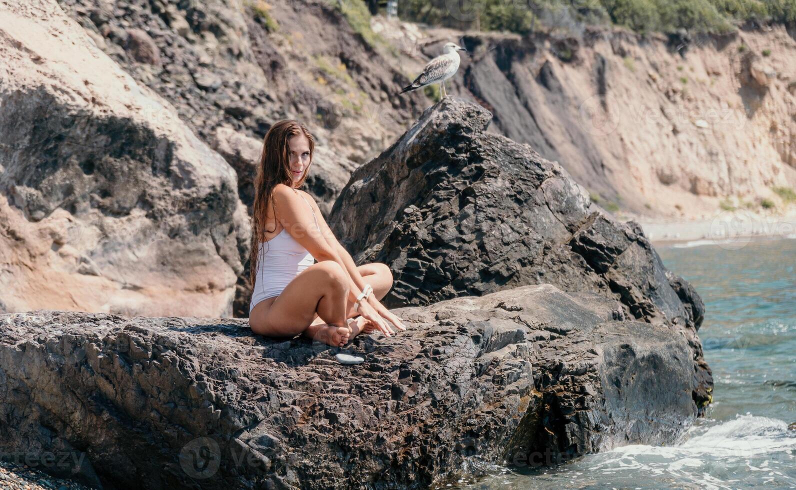mujer viaje mar. contento turista en blanco bikini disfrutar tomando imagen al aire libre para recuerdos. mujer viajero posando en el playa a mar rodeado por volcánico montañas, compartiendo viaje aventuras viaje foto