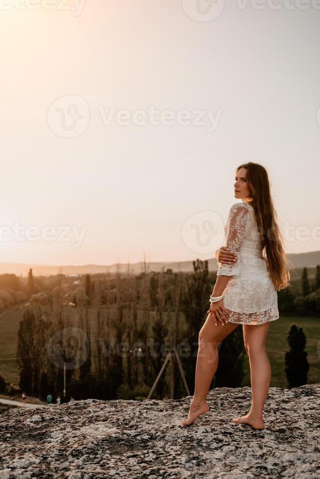 contento mujer en blanco boho vestir en puesta de sol en montañas. romántico mujer con largo pelo en pie con su espalda en el puesta de sol en naturaleza en verano con abierto manos. silueta. naturaleza. puesta de sol. foto