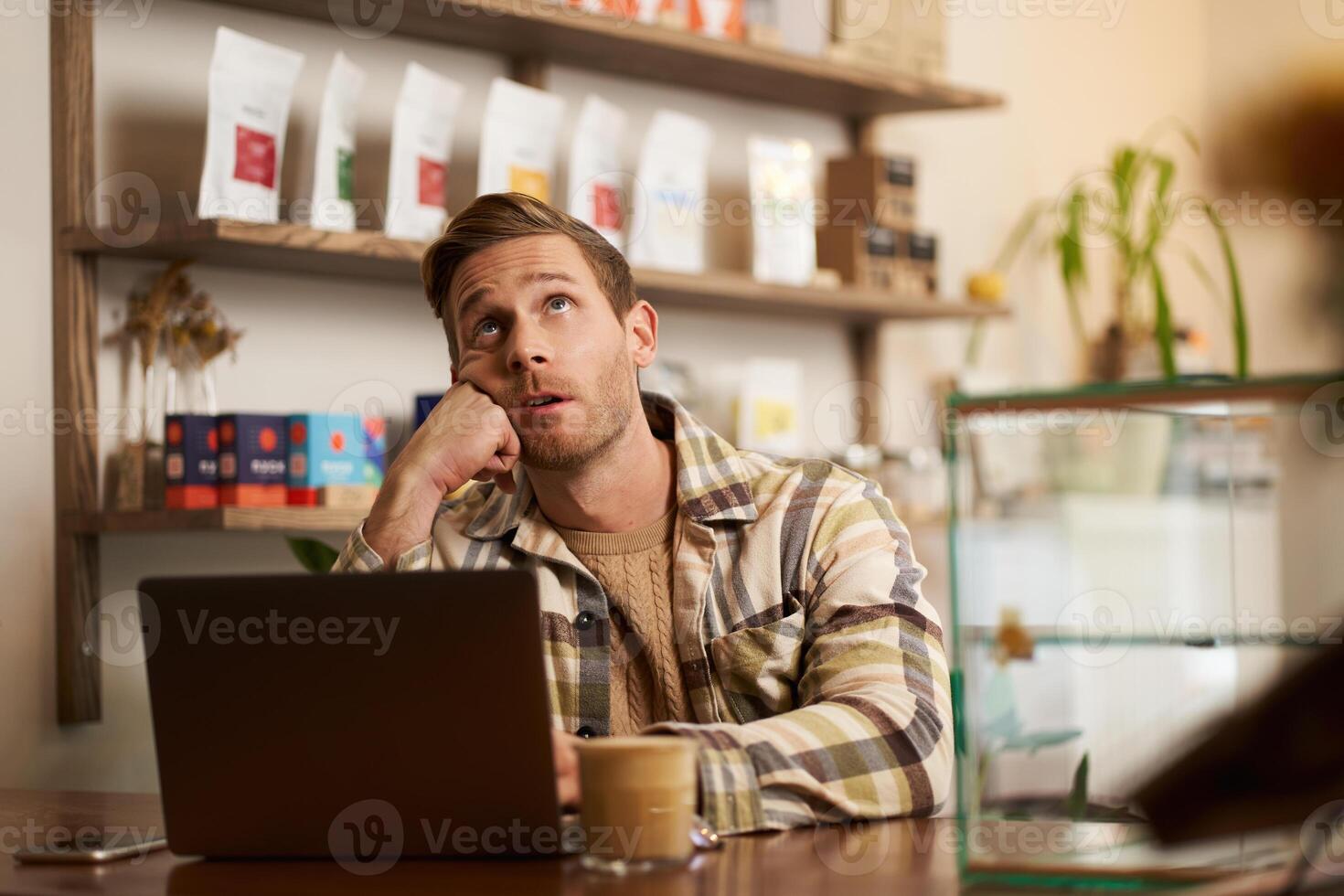 Portrait of stressed working man, sitting in cafe with laptop, business owner doing project online, looking up, roll his eyes, drinking coffee in co-working creative space photo