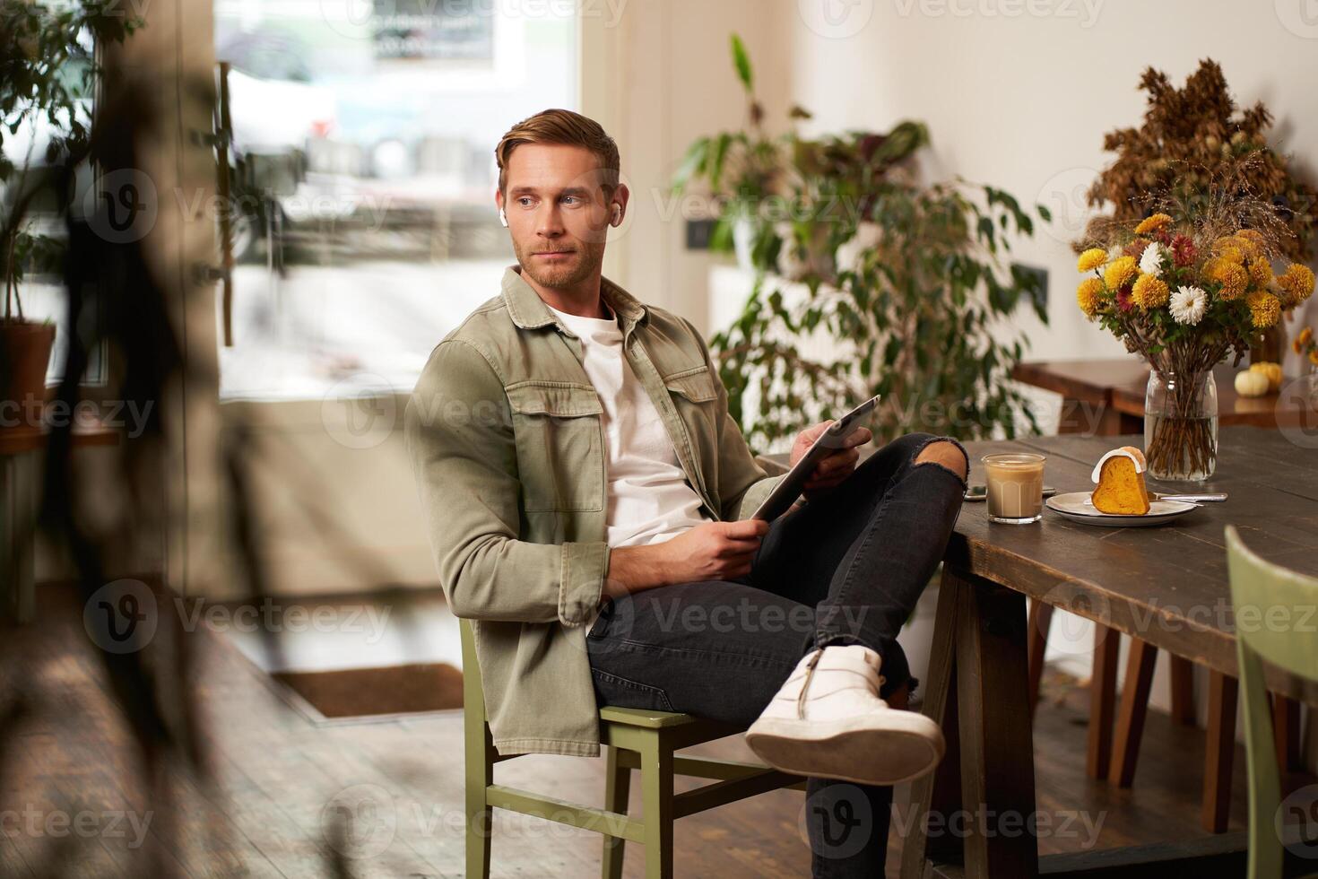 Lifestyle shot of handsome young man sitting in a cafe in front of the table, drinking coffee, wearing wireless headphones, listening to music or watching video in public space, using digital tablet photo