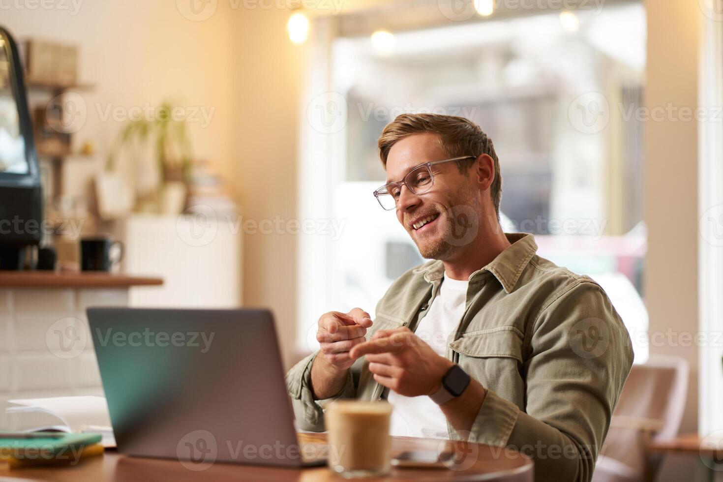 Portrait of handsome, smiling young man, online tutor, businessman working in cafe remotely, waving hand at laptop, connects to video chat or meeting photo