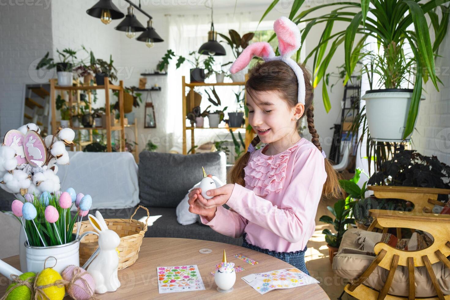 A cute girl with pink bunny ears makes an Easter craft - decorates an egg in the form of a unicorn with rhinestones, horn, flowers in the interior of a house with plants. photo