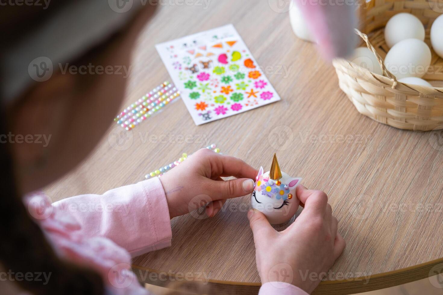 A cute girl with pink bunny ears makes an Easter craft - decorates an egg in the form of a unicorn with rhinestones, horn, flowers in the interior of a house with plants. photo