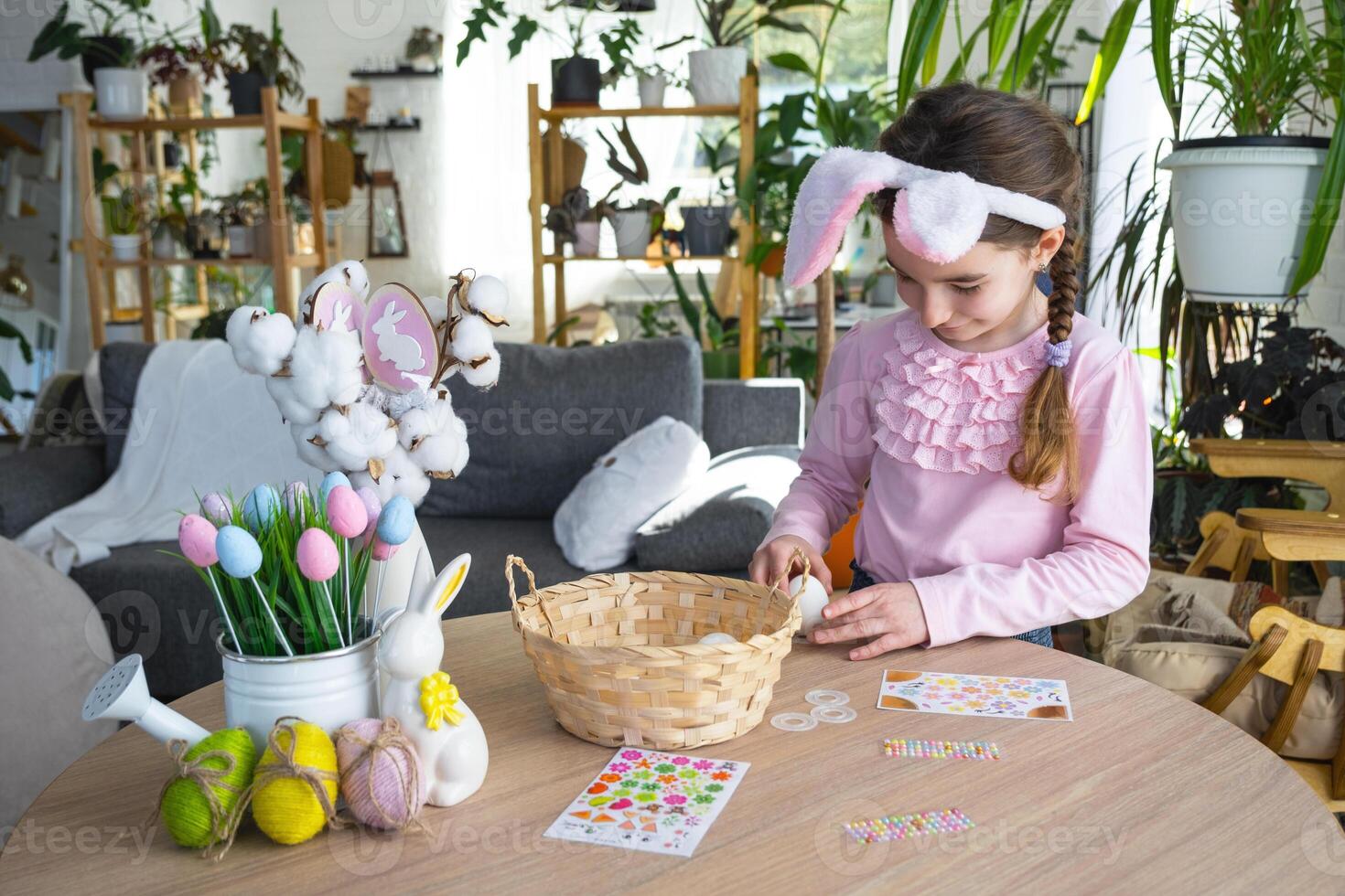 A cute girl with pink bunny ears makes an Easter craft - decorates an egg in the form of a unicorn with rhinestones, horn, flowers in the interior of a house with plants. photo