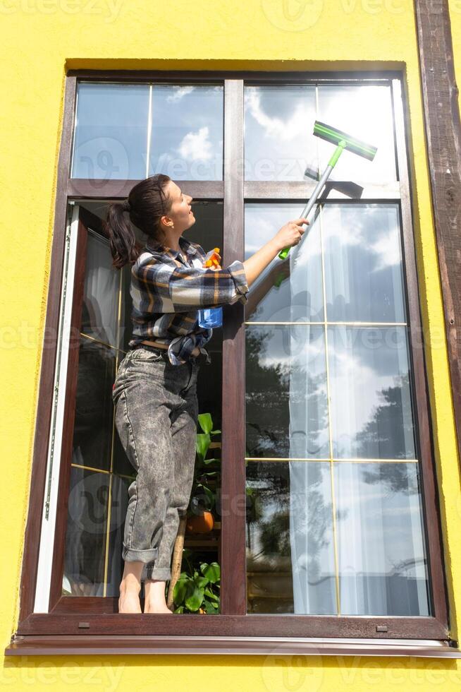 un mujer a mano lavados el ventana de el casa con un trapo con un rociar limpiador y un fregona afuera. la seguridad a altura, restaurar orden y limpieza en el primavera, limpieza Servicio foto