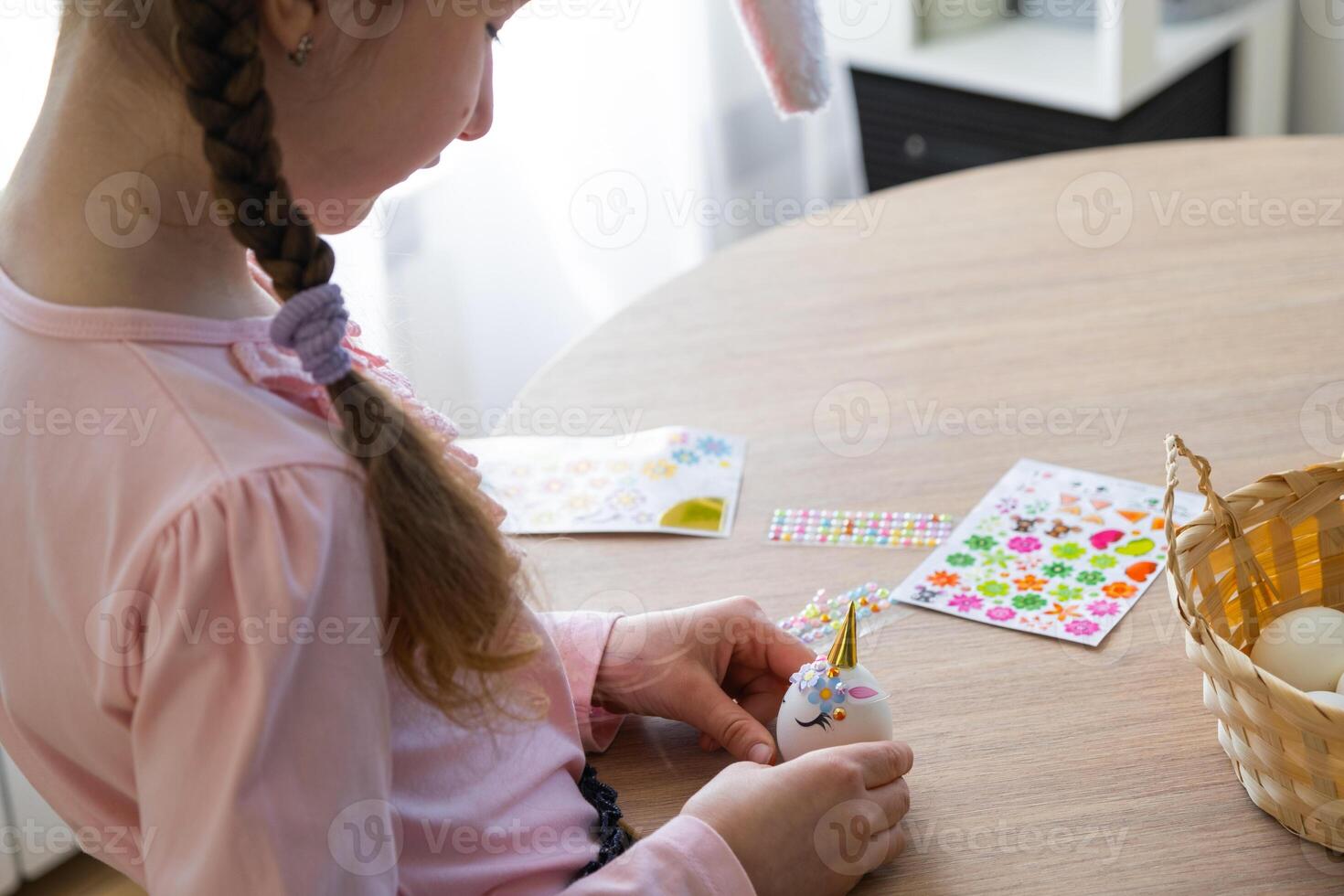 A cute girl with pink bunny ears makes an Easter craft - decorates an egg in the form of a unicorn with rhinestones, horn, flowers in the interior of a house with plants. photo