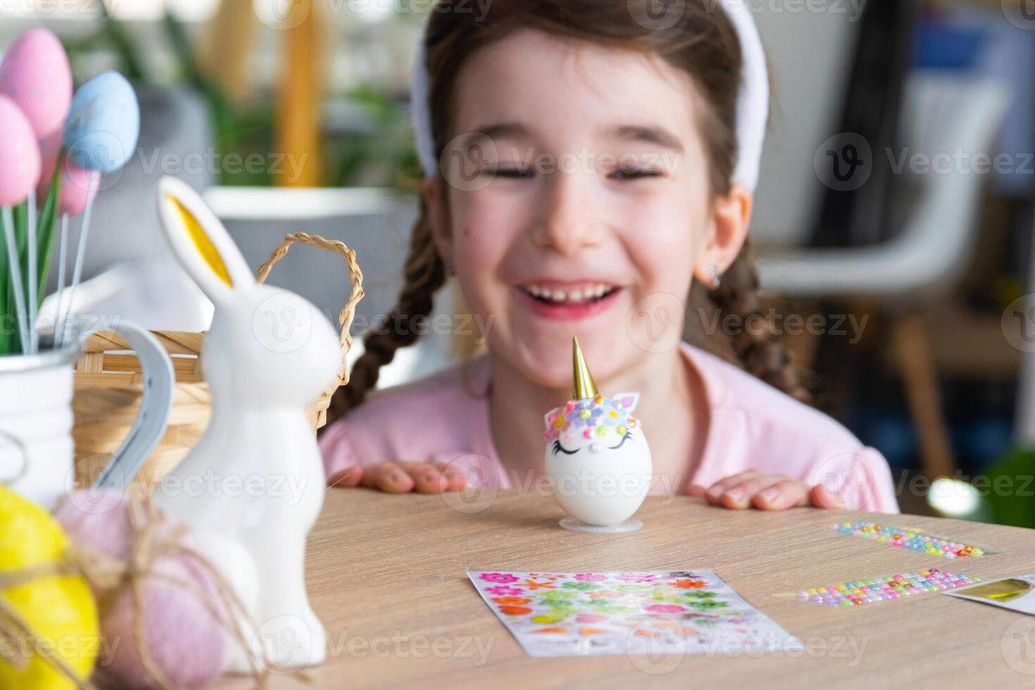 A cute girl with pink bunny ears makes an Easter craft - decorates an egg in the form of a unicorn with rhinestones, horn, flowers in the interior of a house with plants. photo