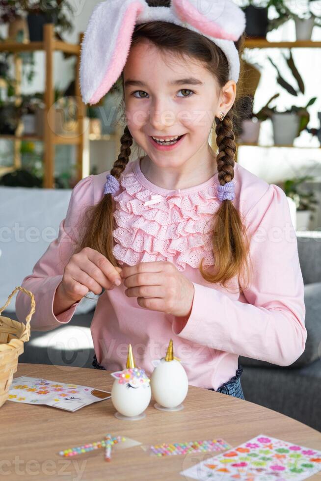 A cute girl with pink bunny ears makes an Easter craft - decorates an egg in the form of a unicorn with rhinestones, horn, flowers in the interior of a house with plants. photo