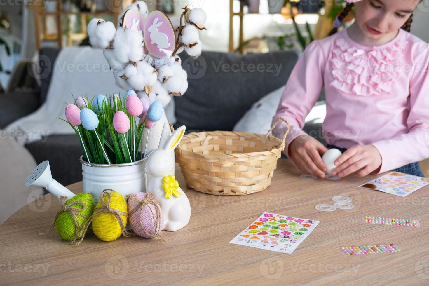 A cute girl with pink bunny ears makes an Easter craft - decorates an egg in the form of a unicorn with rhinestones, horn, flowers in the interior of a house with plants. photo