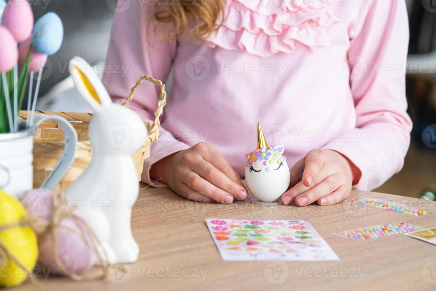 A cute girl with pink bunny ears makes an Easter craft - decorates an egg in the form of a unicorn with rhinestones, horn, flowers in the interior of a house with plants. photo