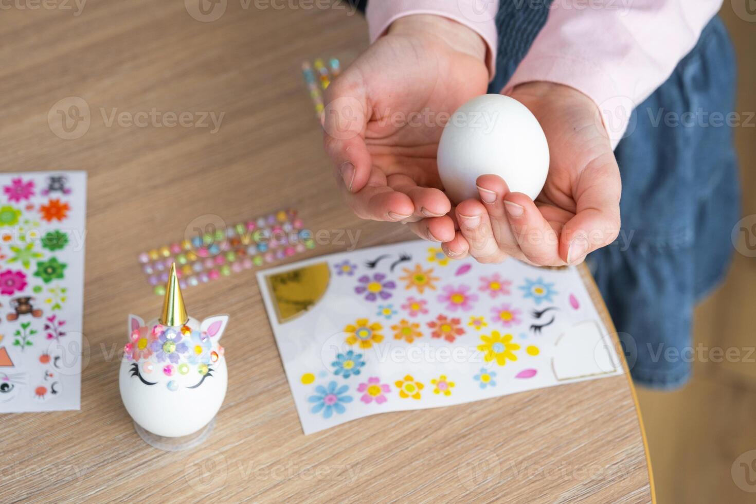 A cute girl with pink bunny ears makes an Easter craft - decorates an egg in the form of a unicorn with rhinestones, horn, flowers in the interior of a house with plants. photo