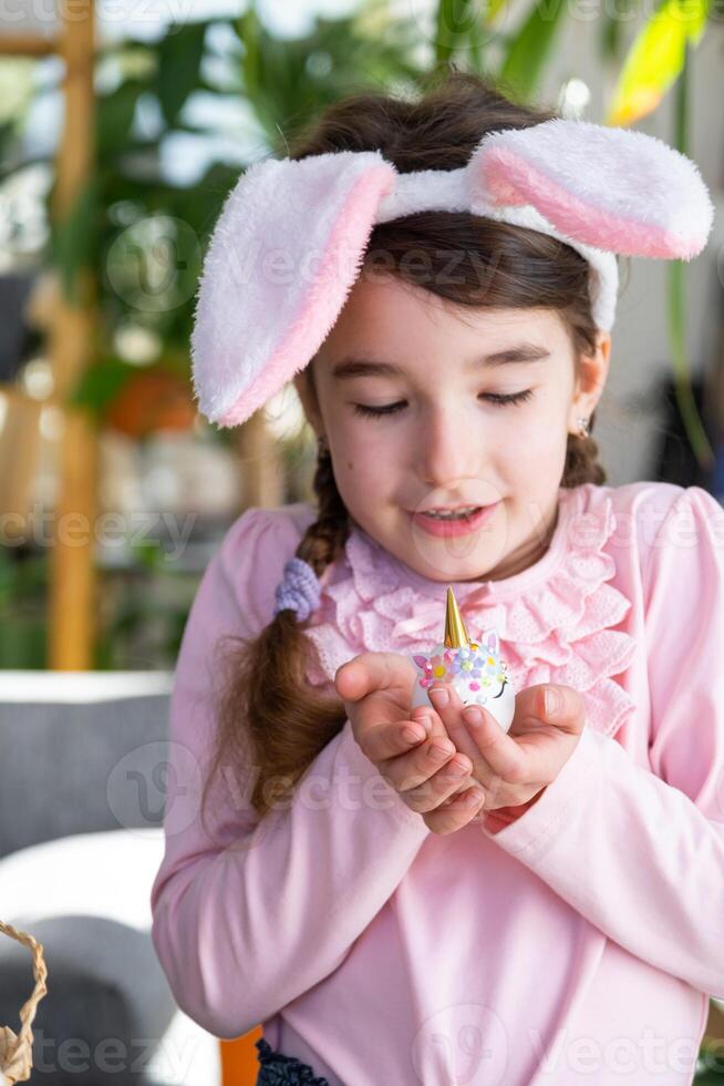 A cute girl with pink bunny ears makes an Easter craft - decorates an egg in the form of a unicorn with rhinestones, horn, flowers in the interior of a house with plants. photo