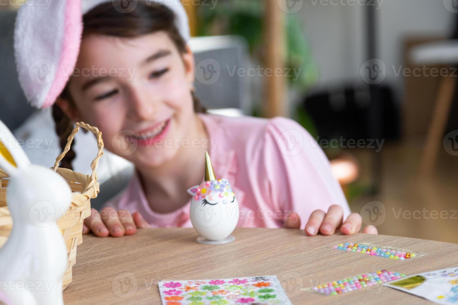 A cute girl with pink bunny ears makes an Easter craft - decorates an egg in the form of a unicorn with rhinestones, horn, flowers in the interior of a house with plants. photo