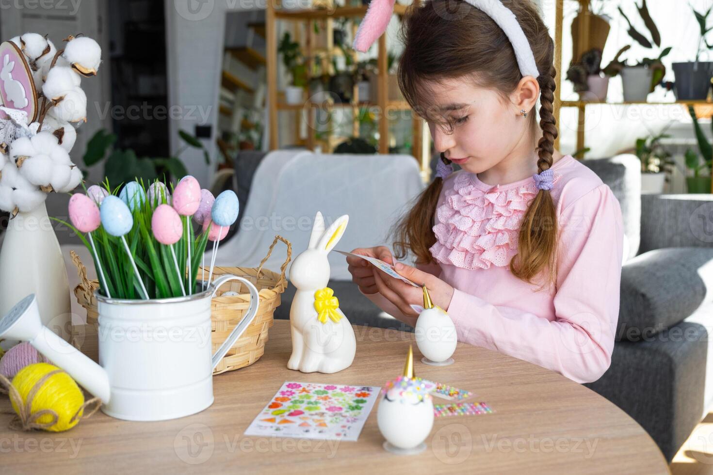 A cute girl with pink bunny ears makes an Easter craft - decorates an egg in the form of a unicorn with rhinestones, horn, flowers in the interior of a house with plants. photo