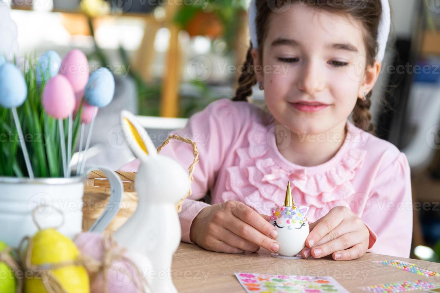 A cute girl with pink bunny ears makes an Easter craft - decorates an egg in the form of a unicorn with rhinestones, horn, flowers in the interior of a house with plants. photo