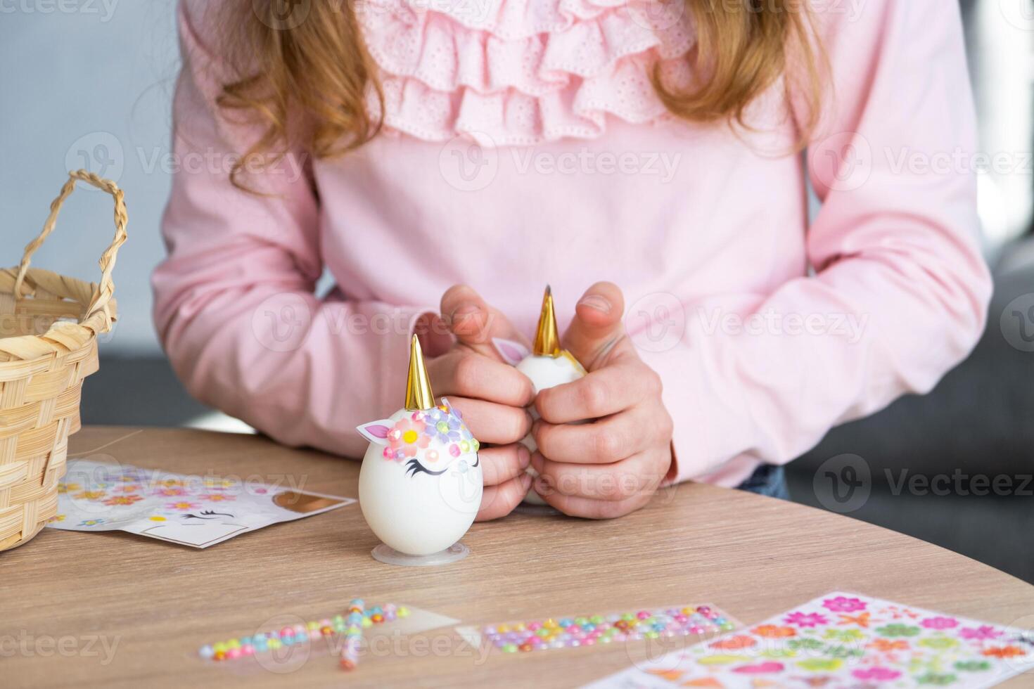 A cute girl with pink bunny ears makes an Easter craft - decorates an egg in the form of a unicorn with rhinestones, horn, flowers in the interior of a house with plants. photo