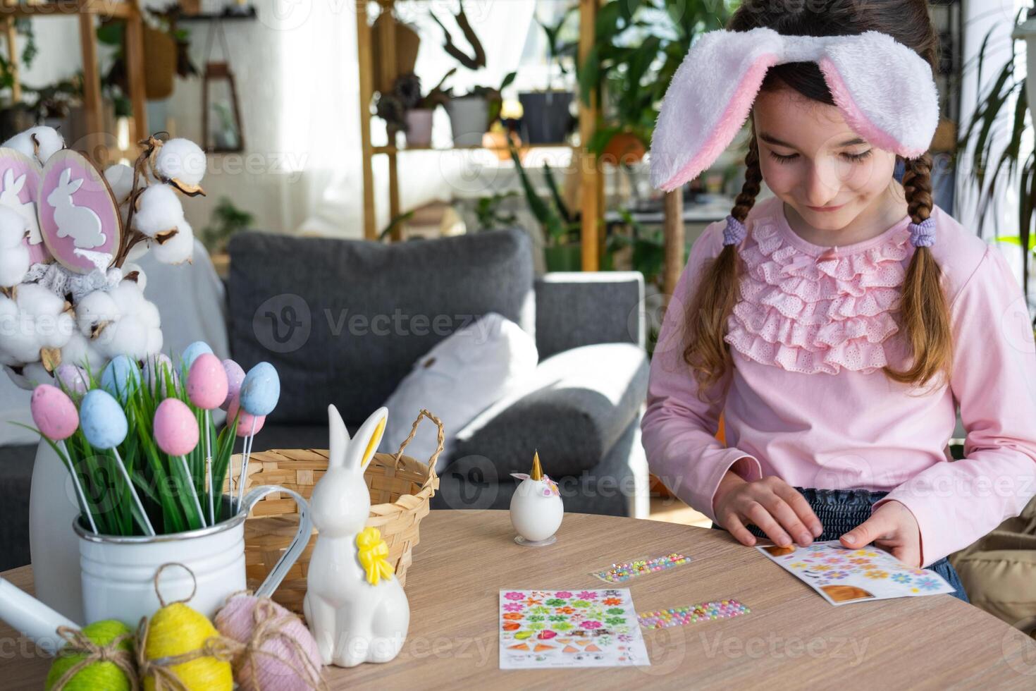 A cute girl with pink bunny ears makes an Easter craft - decorates an egg in the form of a unicorn with rhinestones, horn, flowers in the interior of a house with plants. photo