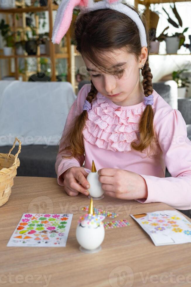 A cute girl with pink bunny ears makes an Easter craft - decorates an egg in the form of a unicorn with rhinestones, horn, flowers in the interior of a house with plants. photo