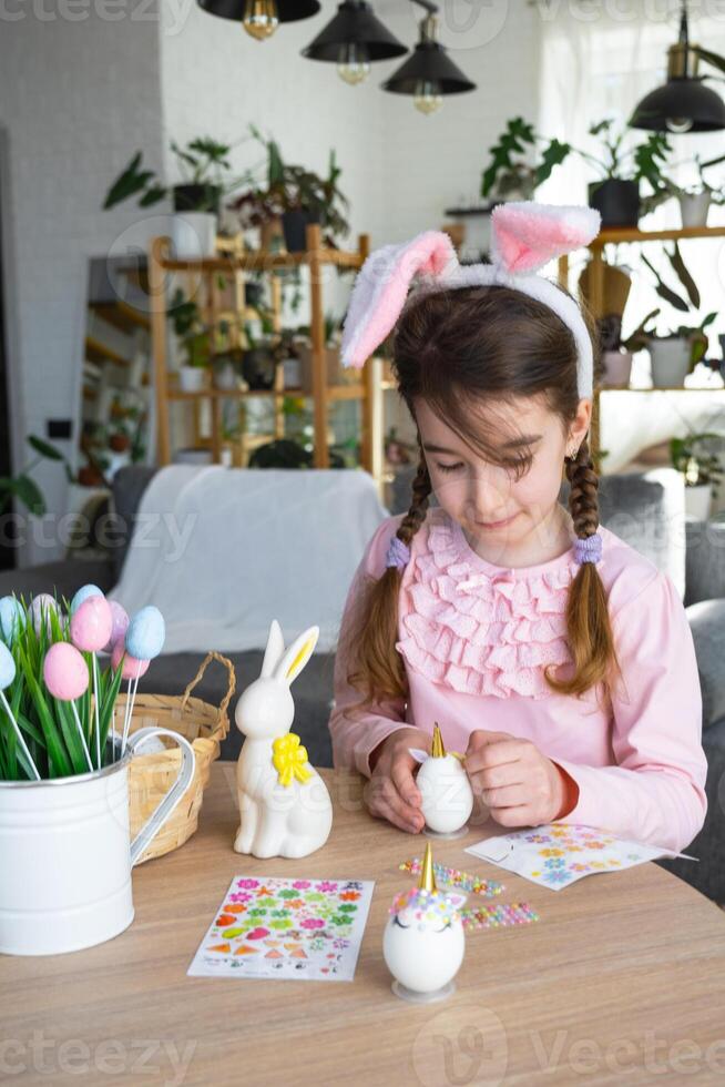 A cute girl with pink bunny ears makes an Easter craft - decorates an egg in the form of a unicorn with rhinestones, horn, flowers in the interior of a house with plants. photo