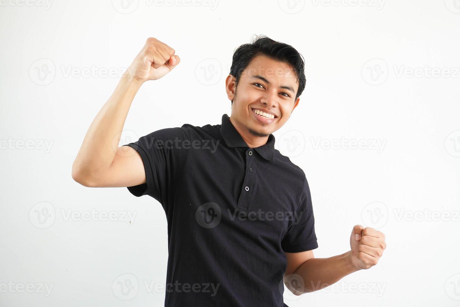 sonriente o contento joven asiático hombre apretado su puños aplausos despreocupado y entusiasmado. victoria concepto, vistiendo negro polo t camisa aislado en blanco antecedentes foto