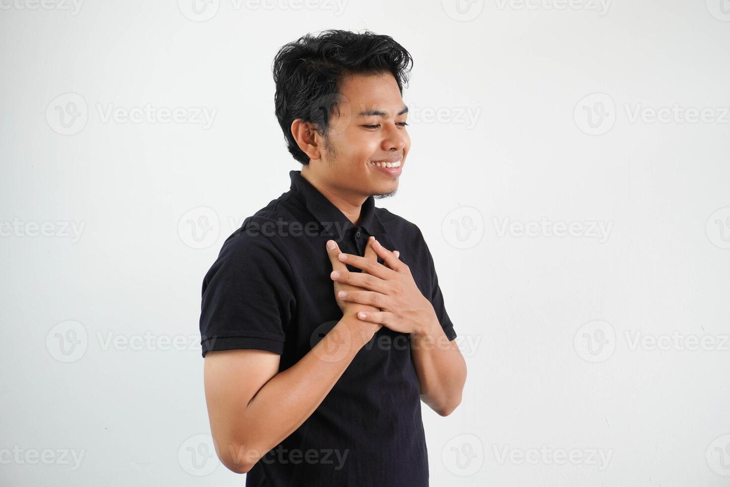 young asian man posing on a white backdrop laughing keeping hands on heart, concept of happiness. wearing black polo t shirt. photo