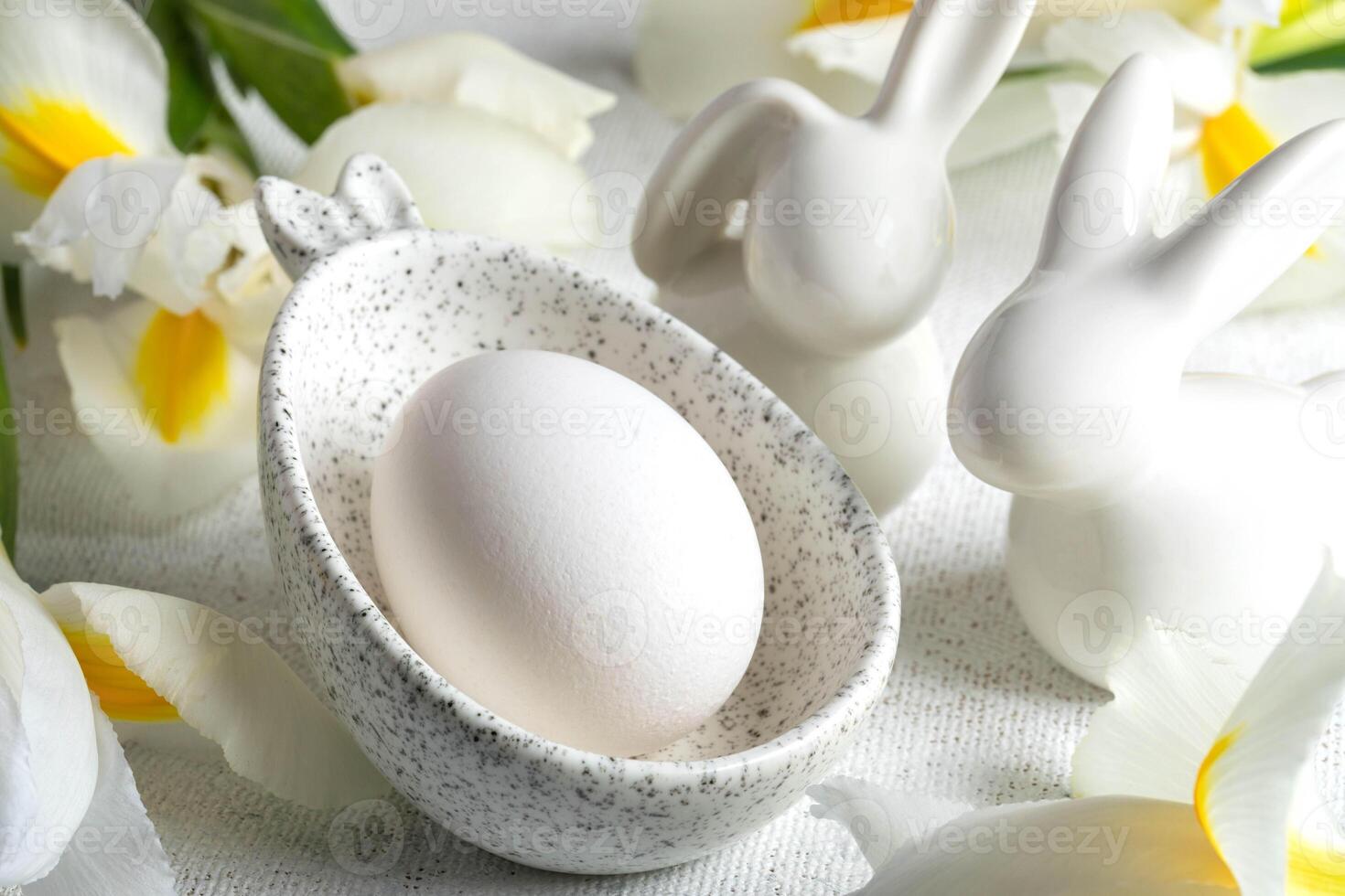 One Easter egg in marble bowl with bunny ears, small bunnies, white Iris flowers on white close up. photo