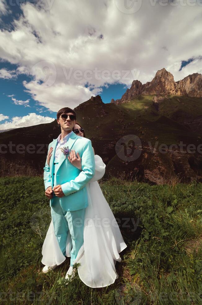 un joven hombre y su esposa estar en un abrazo alto en el montañas en contra el fondo de épico rocas en un soleado día. recién casados Boda Pareja en el montañas foto