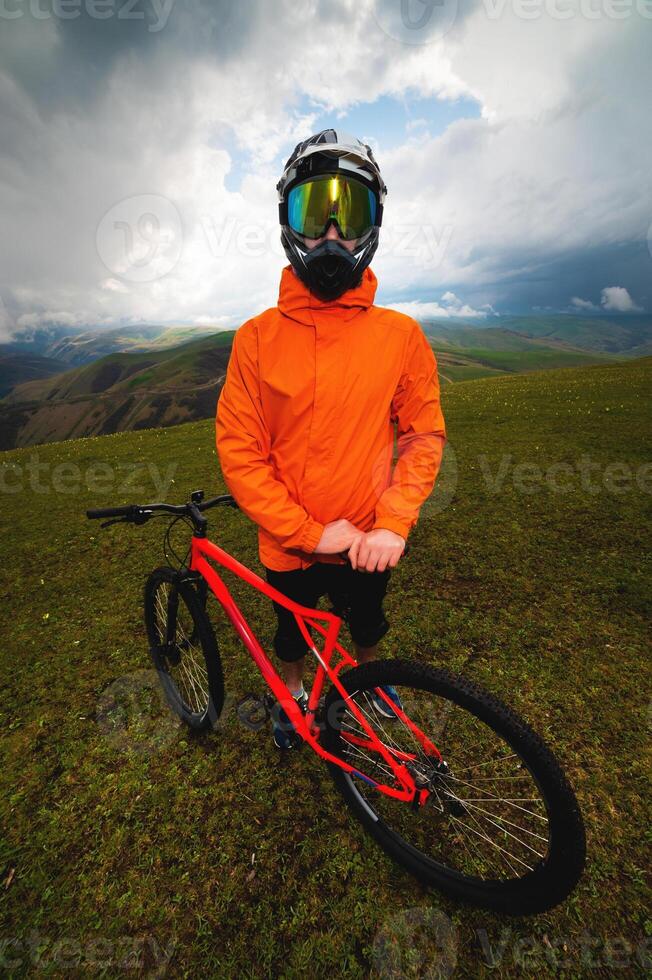 amplio ángulo vertical marco parte superior ángulo. retrato de un barbado montaña motorista con su bicicleta en contra un antecedentes de montañas y nubes en el verano rodeado por verde césped foto