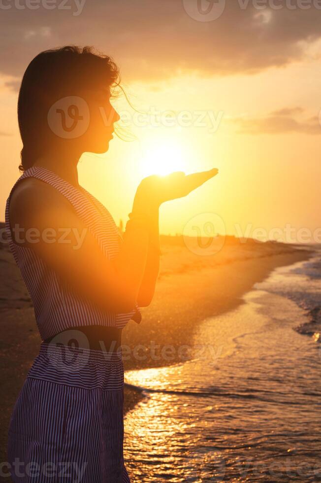 Silhouette of a young Caucasian woman holding the sun in her hands. On the seashore at sunset. photo