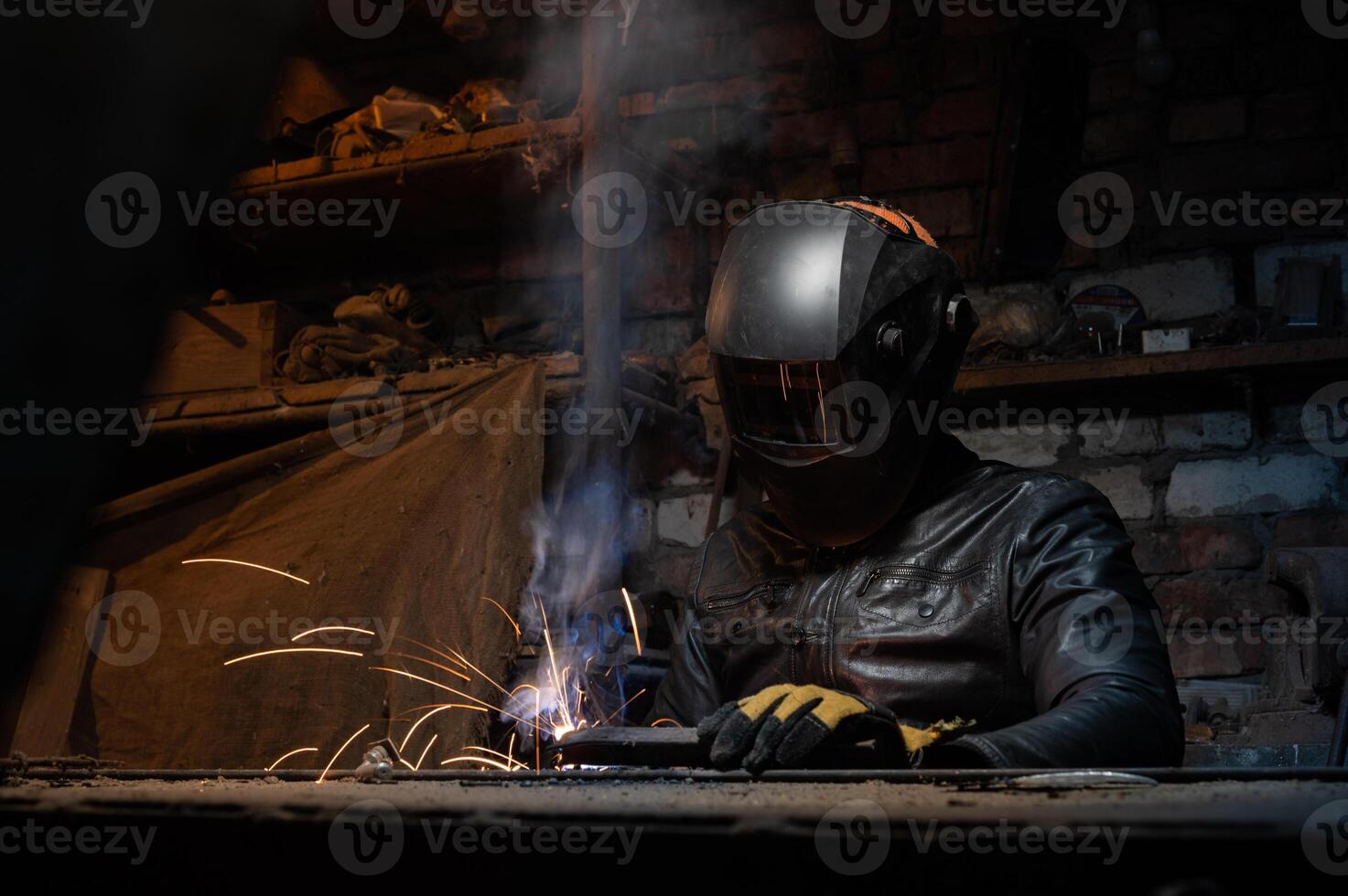 Young mechanic with a welding machine in an old dirty garage at night. Man in protection doing welding work, hobby photo