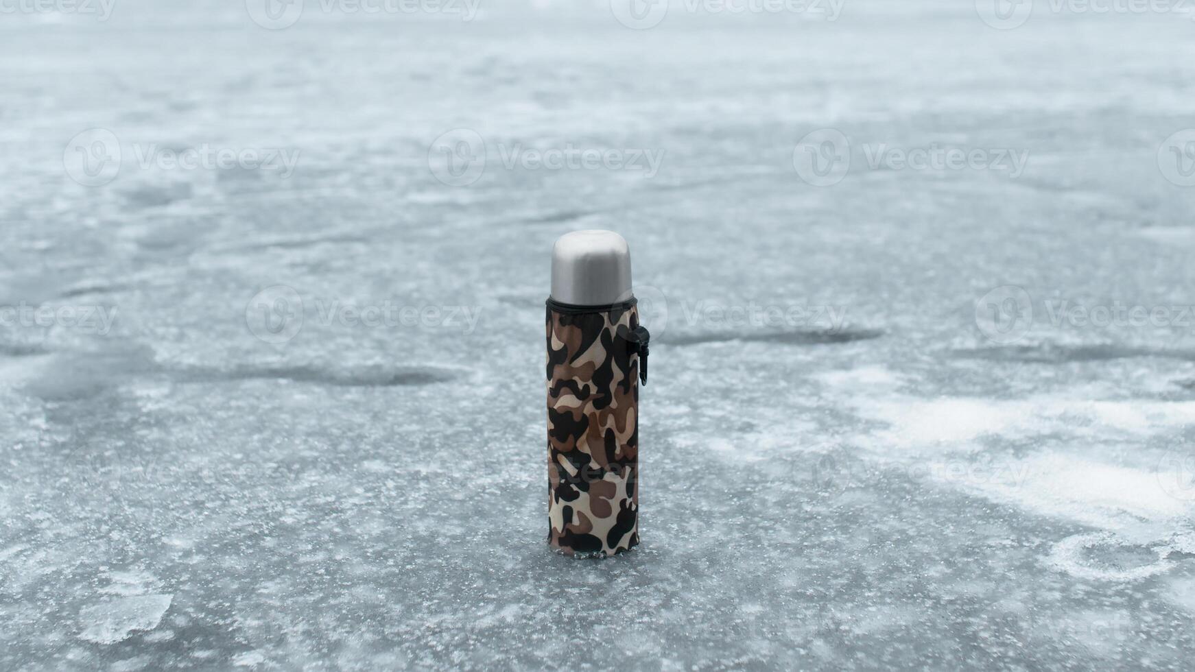 Thermos in a khaki case standing on a frozen lake. Concept hot drink in cold weather, winter fishing, hiking photo