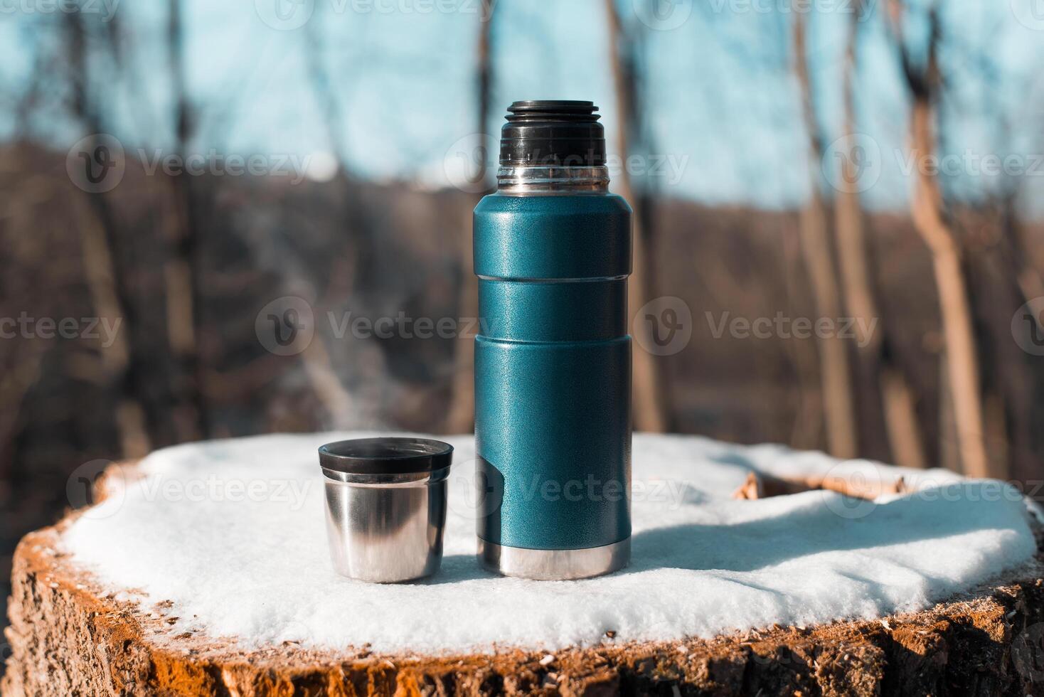 Thermos and mug with hot drink standing on stump. Winter forest, steam from warming drink in a cup. photo