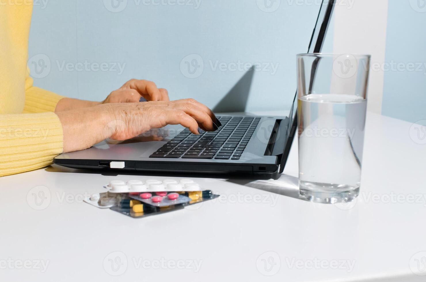 Elderly woman's hands are typing on laptop close-up. Medicines and glass of water standing on table at home. Concept online consultation with doctor, search for instructions on Internet photo